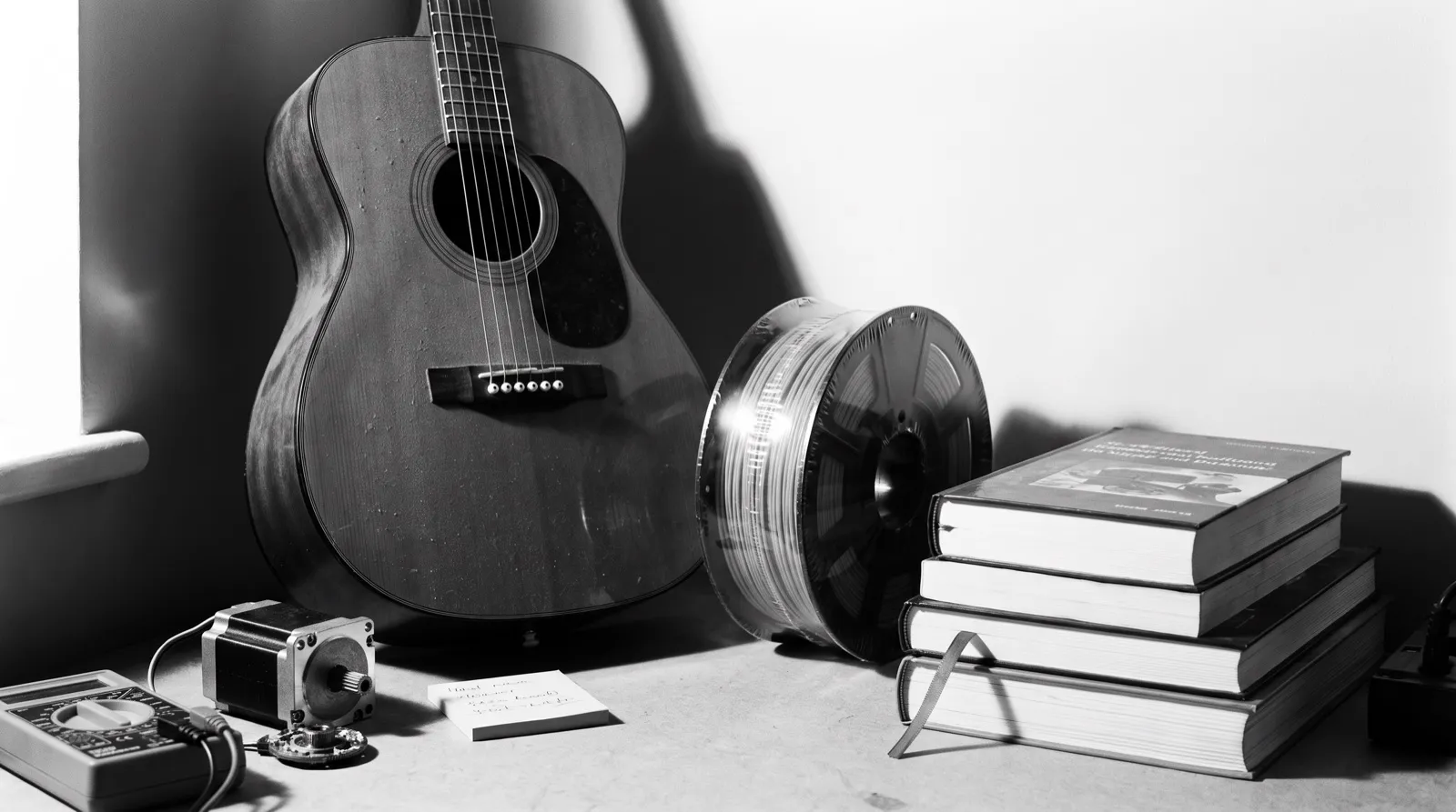 A dim home workbench corner: a dusty guitar, a shrink-wrapped filament spool, a half-disassembled servo motor, a bookmarked technical book